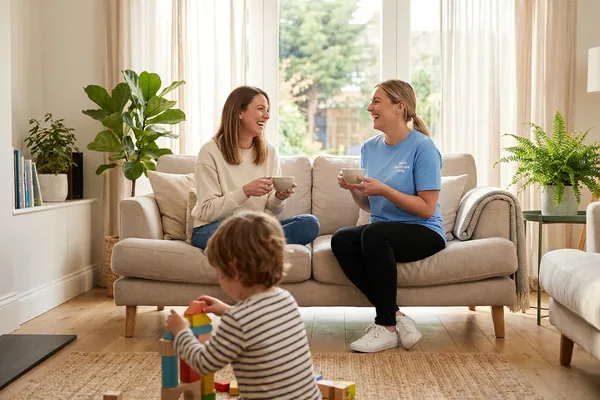 A mother and a Giant Healthcare support worker laughing over tea on a sofa while a young child seen from behind builds with blocks on the rug